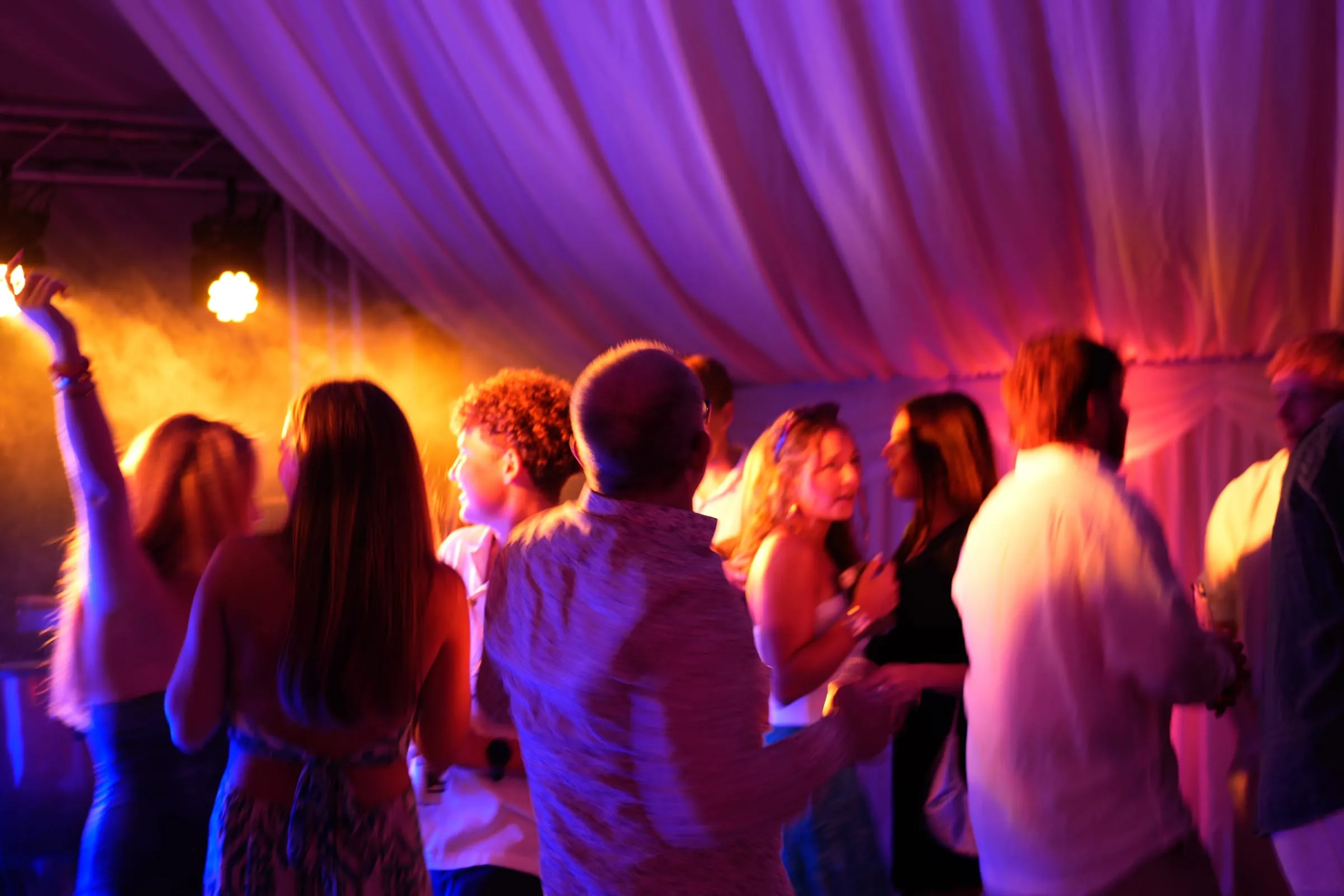 A crowd dancing under orange and violet light, arms raised.