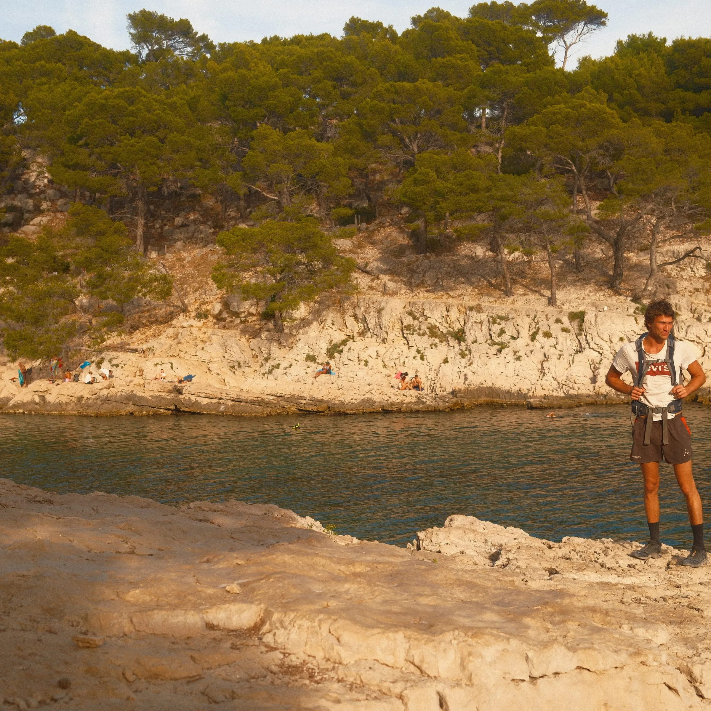 A man stands on the rocks at the edge of a narrow inlet, pines behind him.