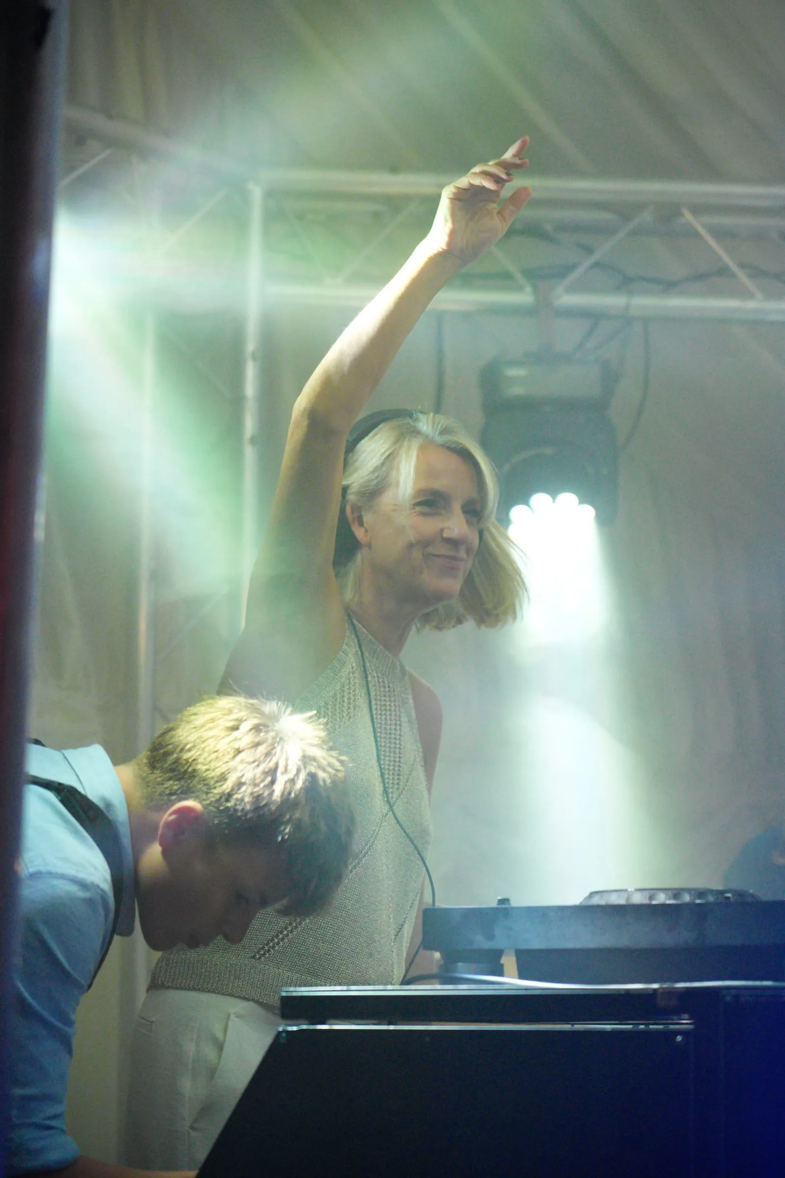 The DJ smiles with a hand in the air, in pale light.